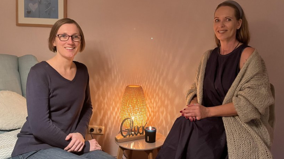 Das Foto zeigt Kerstin Meyer und Conny Köpp, sitzend vor einem kleinen Tisch mit Lampe und einem Deko-Stück mit dem Namen "Glück".